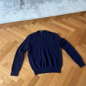 Mörkblå v-ringad tröja från Polo Ralph Lauren - Klassisk mörkblå v-ringad tröja från Polo Ralph Lauren med röd broderad logga på bröstet. Tröjan är stickad i mjukt material och har långa ärmar samt ribbade muddar vid ärmslut och nederkant. Perfekt för lager-på-lager-stil.