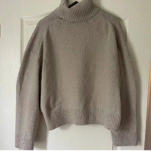 Beige stickad polotröja med ribbad krage - Snygg beige polotröja med bred ribbad krage och muddar. Tröjan är stickad och har en loose passform med långa ärmar. Perfekt för att hålla sig varm och se chill ut under höst och vinter. Enkel att matcha med jeans eller mjukis.