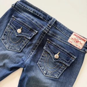 True Religion Joey Low Rise Flare Jeans - Säljer ett par blå True Religion Joey jeans med låg midja och flare-ben. Klassiska kontrastsömmar, stora fickor med lock och silverfärgade knappar. Snyggt broderad logga på bakfickorna och patch bak i midjan. Tillverkade i jeansmaterial. I jätte fint skick! Enda stället det synts att de använts är insidan av ena benets nederkant , se bild. 