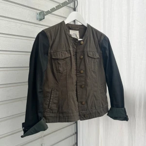 Olivgrön jacka med svarta ärmar H&M - Snygg olivgrön jacka från H&M L.O.G.G. med svarta ärmar i kontrastmaterial. Jackan har knappar framtill, två bröstfickor med lock och raka linjer.