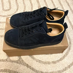 Louboutin skor - Snygga svarta sneakers från Louboutin med klassisk röd sula och diskret logga på sidan. Ovandel i mocka och insida i beige skinn ger en lyxig känsla. Skorna har rund tå, svarta snören och platt sula. Perfekta för dig som gillar stilrena och exklusiva sneakers.