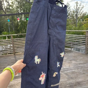 Blå täckbyxor med enhörningspatches H&M - Mörkblå täckbyxor från H&M med elastisk midja och flera färgglada enhörningspatches och regnbåge på benen. Byxorna är fodrade och har en praktisk knappstängning framtill. Perfekta för kalla dagar när du vill sticka ut lite extra.