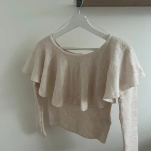 Beige stickad tröja med volang H&M - Superfin beige stickad tröja från H&M i storlek S. Tröjan har en bred volang över axlarna och är långärmad med lite off shoulder.Perfekt för dig som gillar en söt och trendig look med extra detalj vid halsringningen.