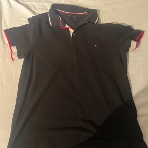 Svart piké från Tommy Hilfiger - Svart pikétröja från Tommy Hilfiger med klassisk krage och smal passform. Detaljer i vitt och rött på krage och ärmslut samt liten broderad logga på bröstet. Perfekt för en clean och stilren look.