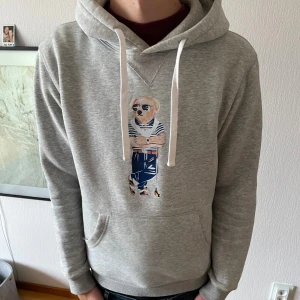 Grå hoodie Polo Bear från Ralph Lauren - Säljer en grå hoodie från Ralph Lauren med Polo Bear-tryck på bröstet. Tröjan har vit snörning i huvan, känguruficka och ribbade muddar. Skön och stilren, perfekt för dig som gillar klassiska streetwear-vibbar.