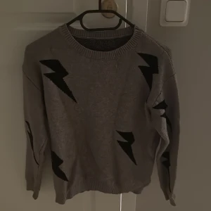 Grå stickad tröja med blixtar - Grå stickad tröja med svarta blixtmönster över hela tröjan. Rund halsringning och långa ärmar med ribbade muddar. Perfekt för dig som gillar en cool och unik stil.