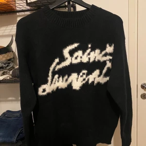Svart stickad Saint Laurent-tröja - Säljer en svart stickad tröja från Saint Laurent med vit, fluffig logga framtill. Tröjan har rund halsringning och långa ärmar. Den är tillverkad i Italien och har en oversized vibe med riktigt mjuk känsla. Perfekt för dig som vill sticka ut med en lyxig statement-piece.