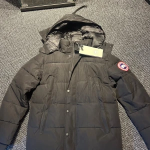Svart jacka från Canada Goose - Svart dunjacka från Canada Goose med huva och klassisk logopatch på ärmen. Jackan har quiltad design, dragkedja och knappar framtill samt två sidofickor. Perfekt för kalla dagar och har en stilren look som funkar till det mesta.