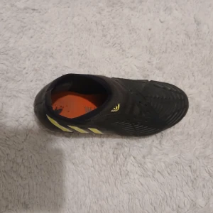 Adidas Predator svarta fotbollsskor - Adidas Predator fotbollsskor i svart med gula och vita detaljer. Skorna har en strömlinjeformad design utan snörning, med mönstrad ovandel för extra grepp på bollen. De klassiska tre ränderna på sidan och Adidas-logga på tungan. Snygg och modern look för dig som gillar att sticka ut på planen.