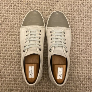 Lanvin sneakers med grå lacktå - Snygga sneakers från Lanvin i ljusbeige mocka med grå lackad rund tå. Skorna har vita snören och metallfärgade öljetter, samt en platt sula i matchande ton. Perfekt för dig som gillar stilrena och exklusiva detaljer. Mockan är en blandning mellan vit och beige väldigt snygg! Fråga på så mycket som helst!