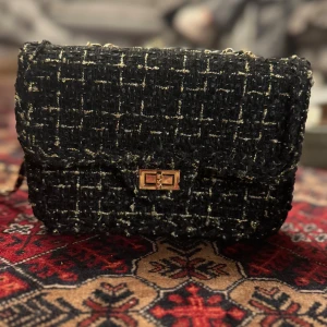  axelväska med kedja - Snygg svart axelväska med rutigt mönster i guldton och grov textur. Väskan har en guldfärgad kedja som axelrem och ett elegant vridlås i guld på framsidan. Perfekt storlek för det viktigaste och ger en cool vibe till din outfit.