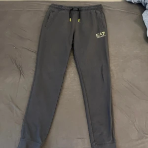 Svarta mjukisbyxor EA7 Emporio Armani - Svarta mjukisbyxor från EA7 Emporio Armani med dragsko i midjan och gul logga på ena benet. Byxorna har smal passform, ribbade muddar vid benslut och tryckdetaljer på baksidan av benet. Perfekta för chill eller träning.