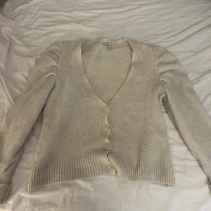 Stickad kofta  - Mysig beige kofta från Stradivarius i storlek S. Koftan har v-ringning, ribbade muddar och fyra knappar framtill. Långärmad med lätt puff vid axlarna för en trendig touch. Perfekt att slänga över en t-shirt eller linne.