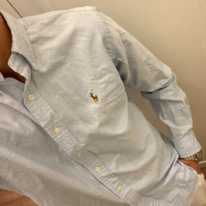 Ljusblå Skjorta från Polo Ralph Lauren (Slim Fit) - Snygg ljusblå skjorta från Ralph Lauren i slim fit med klassisk button-down krage och broderad logga på bröstet. Skjortan har långa ärmar, vita knappar och är tillverkad i mjuk bomull. Perfekt för dig som gillar stilrena och tidlösa plagg. Storlek Medium. Snabb Leverans!