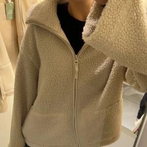 Beige teddyjacka med dragkedja - Mysig beige teddyjacka med hög krage och två stora fickor framtill. Jackan har en hel dragkedja och är tillverkad i mjukt fleece-material som ger en fluffig och chill look. Perfekt för kalla dagar när du vill vara både varm och snygg.