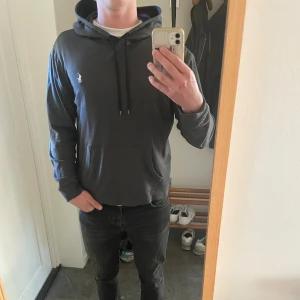 Mörkgrå hoodie från Ralph Lauren - Säljer en mörkgrå hoodie från Ralph Lauren med vit broderad logga på bröstet. Tröjan har huva med snörning, magficka och långa ärmar. Perfekt för chill dagar och enkel att matcha med jeans eller joggers.