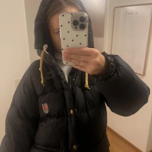 Svart pufferjacka från Fjällräven - Säljer en svart pufferjacka från Fjällräven med gul snörning i huvan och klassisk logga på bröstet. Jackan har huva, dragkedja och knappar framtill, samt en oversized passform som är perfekt för kalla dagar. Har defekter då en knapp saknas och använd men är i fuktion!!