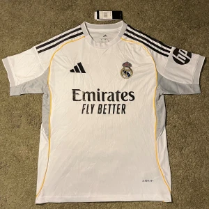 Real Madrid matchtröja Adidas vit - Säljer en vit Real Madrid matchtröja från Adidas med svarta och gula detaljer. Tröjan har klubbmärke och sponsortryck på bröstet samt svarta ränder på axlarna. Tillverkad i lätt och ventilerande material, perfekt för fotboll eller träning.