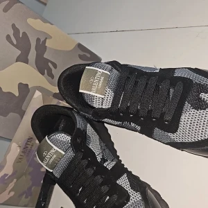 Valentino Garavani Rockrunner sneakers - Snygga Valentino Garavani Rockrunner sneakers i svart, grått och silver med camo-mönster 🫟. Skorna har mesh och mockadetaljer, svarta snören och en robust sula med coola nitar. Size 44 men passar också 43😇. Perfekta för dig som vill sticka ut med exklusiva och trendiga sneakers💫