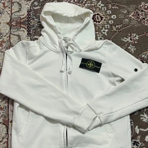 Vit hoodie från Stone Island - Vit hoodie från Stone Island med klassisk logotyp på ärmen. Tröjan har huva med snörning, dragkedja framtill och ribbade muddar. Perfekt för dig som gillar streetwear och vill ha en clean look.