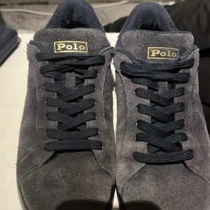 Mörkblåa sneakers från Polo Ralph Lauren - Snygga mörkblåa sneakers från Polo med ovandel i mocka. Skorna har guldiga Polo-loggor både på plösen och hälen. Klassisk låg modell med platt sula och rund tå, perfekt för en stilren och avslappnad look. Färgen lite mer blekt än original men går att putsa upp mockan. 
