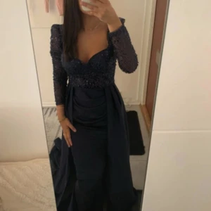 Marinblå långklänning med paljetter - Säljer en marinblå långklänning med glittrig paljettöverdel och djup v-ringning. Klänningen har långa ärmar i transparent tyg och ett svepande, elegant kjolparti som ger en riktigt lyxig känsla. Perfekt för bal eller fest! Kjolen är i mjukt, flowy material.