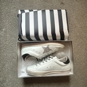 Golden Goose vita sneakers med glitterstjärna - Säljer ett par vita Golden Goose sneakers med silverglittrig stjärna på sidan, grå snörning och svart detalj på hälen. Skorna har platt sula och är tillverkade i skinn för en clean och trendig look. Perfekta för dig som vill sticka ut med lite extra bling.