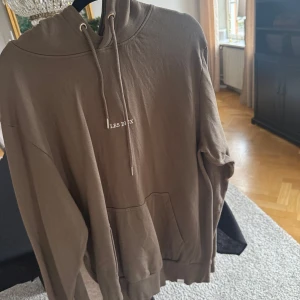 Brun hoodie från Les Deux med snörning - Säljer en brun hoodie från Les Deux med broderad logga på bröstet. Tröjan har huva med snörning, känguruficka och ribbade muddar. Perfekt för chill dagar och enkel att matcha med jeans eller joggers.