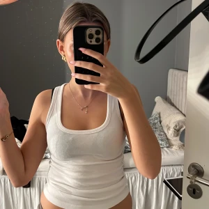 Vit ribbad linnetopp - Säljer en klassisk vit ribbad linnetopp med breda axelband och rundad urringning. Perfekt att styla till jeans eller kjol för en enkel och fräsch look. Materialet är mjukt och stretchigt för en bekväm passform.