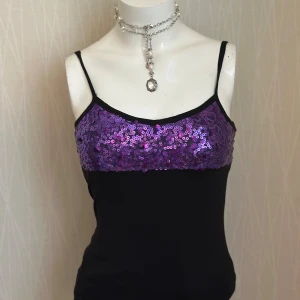 Svart linne med lila paljetter - Svart linne från Bik Bok med smala axelband och lila paljetter över bysten. Toppen har en snygg öppen rygg med korsade band och paljettpanel även bak. Materialet är mjukt och stretchigt, perfekt för att sticka ut med glittriga detaljer.