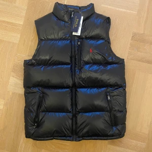 Svart dunväst från Polo Ralph Lauren - Snygg svart dunväst från Polo Ralph Lauren med hög krage och två praktiska fickor med dragkedja. Västen har ett quiltat mönster och den klassiska röda Polo-loggan på bröstet. Perfekt att slänga över en hoodie nu innan hösten! Västen är i XL - 18-20 år!!!