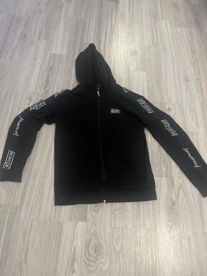 Svart EA7 hoodie med logga och dragkedja - Svart hoodie från EA7 Emporio Armani med hel dragkedja och huva. Snygga logotyper längs ärmarna och på bröstet, samt coola gula detaljer vid dragkedjan. Tillverkad i mjuk bomull för en skön känsla. Perfekt för en streetwear-look.            MÖTS I STOCKHOLM🙃