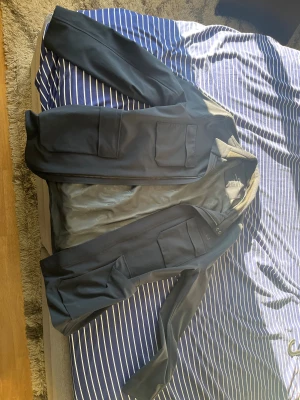 Svart jacka från UBR, X-Large - Snygg svart jacka från UBR i storlek X-Large. Jackan har flera praktiska fickor framtill, dragkedja och en stilren design. Perfekt för dig som gillar minimalistisk och funktionell stil. Materialet känns tekniskt och lätt, med en modern passform.