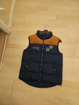 Blå dunväst med bruna detaljer Lindex - Snygg blå dunväst från Lindex med bruna mockadetaljer över axlarna och broderade märken på bröstet. Västen har dragkedja framtill, två fickor med tryckknappar och quiltad design. Perfekt för att addera lite extra stil till din outfit.