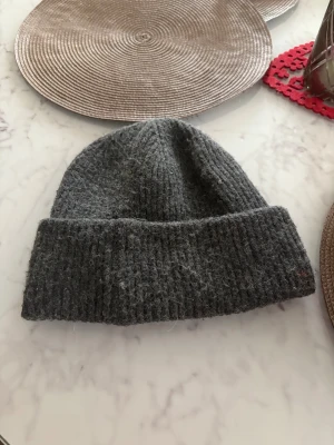 Grå ribbad ullmössa från lager 157 - Snygg grå mössa från lager 157 i ribbstickad ull. Klassisk form med uppvikt kant och grovstickad struktur som ger en chill vibe. Perfekt för kalla dagar och enkel att matcha med din jacka eller hoodie.
