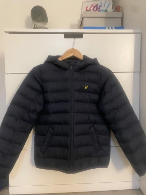 Mörk blå pufferjacka från Lyle & Scott - Svart pufferjacka från Lyle & Scott med huva och dragkedja framtill. Jackan har det klassiska gula fågel-loggan på bröstet och är vadderad för extra värme. Perfekt för kyliga dagar och har en stilren, enkel look.