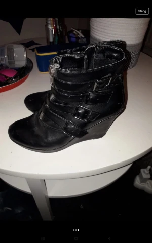 Svarta stövletter med spännen och klack - Säljer ett par svarta stövletter i skinnimitation med flera dekorativa spännen och dragkedja framtill. Skorna har kilklack och rund tå, vilket ger en edgy och cool vibe. Perfekta för dig som vill sticka ut med din stil.