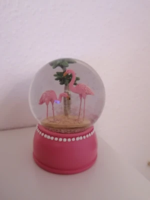 En rosa snöglob med två flamingos  - Denna produkt är en dekorativ snöglob med två rosa flamingos och ett palmträd inuti, placerad på en rosa fot med vita pärldetaljer. Perfekt som prydnad eller present. Inga defekter eller slitage. Ej elektronik.