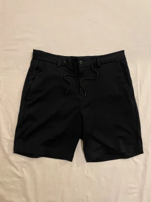 Svarta shorts från Selected Homme - Svarta shorts från Selected Homme med snörning i midjan, knappstängning och bälteshällor. Klassisk rak modell med fickor fram och bak, samt elastisk del baktill i midjan. Perfekta för varma dagar och stilrena outfits.