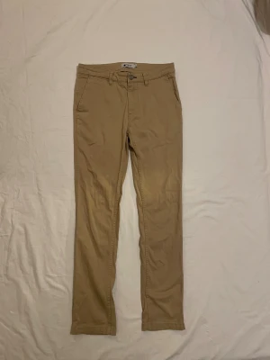 Beiga chinos från NN07 - Snygga beiga chinos från NN07 med klassisk rak passform och midwaist. Byxorna har framfickor, bakfickor med knapp och bälteshällor. Tillverkade i mjuk bomull som är skön att ha på sig hela dagen. Perfekt för dig som gillar stilrena och enkla byxor. W29 L 32
