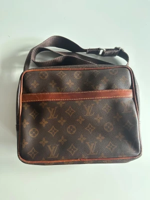 Louis Vuitton vintage bag - Snygg brun axelväska från Louis Vuitton med klassiskt monogrammönster och detaljer i skinn. Väskan har två stora fack med dragkedja, justerbar axelrem och guldfärgade metalldetaljer. Perfekt för dig som vill ha en ikonisk designeraccessoar.