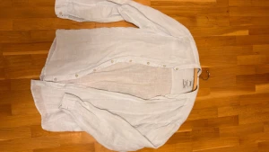 Ljusblå linneskjorta från Pull&Bear - Luftig och stilren ljusblå skjorta från Pull&Bear i 100% linne. Skjortan har klassisk krage, långa ärmar och knäppning framtill. Perfekt för en fräsch och avslappnad look under varma dagar.