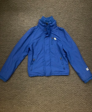 Blå vindjacka från Abercrombie & Fitch - Blå vindjacka från Abercrombie & Fitch med gul broderad älg på bröstet och A&F 1892 på ärmen. Jackan har hög krage, ribbade muddar, flera dragkedjefickor och snygga detaljer i gult. Perfekt för kyliga dagar och enkel att matcha.