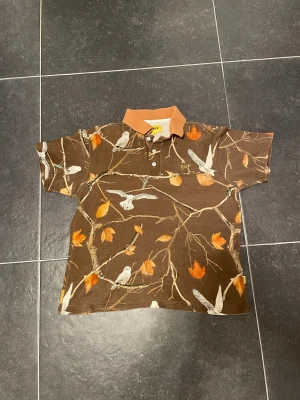 Golf Wang brun camo polo S - Brun kortärmad polo från Golf Wang med camouflagemönster av grenar, fåglar och orange höstlöv. Kragen är i kontrasterande ljusbrun färg och skjortan har knappar framtill. Tillverkad i 100% bomull för en skön känsla.