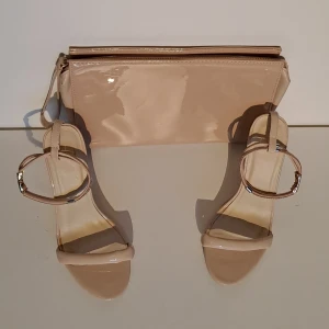 Beige sandaler med glitterdetalj och väska - Snygga beige sandaler med smal hög klack, ca 8 cm. Remmarna har glittriga detaljer och ett spänne i guld. Skorna har öppen tå och platt sula framtill. Materialet är blankt syntet som ger en lyxig känsla. Perfekt för dig som gillar stilrena och glittriga skor. Väska är i liknande nyans