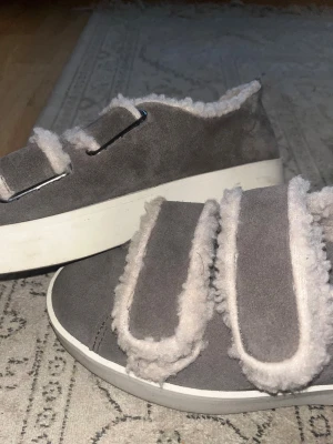 Grå sneakers med fuskpäls från Duffy - Grå sneakers från Duffy med fluffig fuskpäls på insidan och längs kardborrebanden. Skorna har en platt vit sula och är tillverkade i mocka-liknande material. Rund tå och dubbla kardborreband ger en chill och mysig vibe, perfekt för kalla dagar.