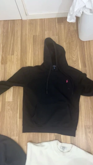 Svart hoodie från Polo Ralph Lauren - Svart hoodie från Polo Ralph Lauren med klassisk huva och snörning. Tröjan har en rosa broderad logga på bröstet och en stor magficka framtill. Perfekt för dig som gillar stilrena och bekväma plagg med en sportig touch.