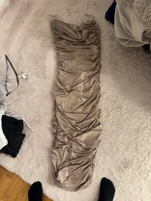 Beige långklänning med rynkade detaljer - Supersnygg beige långklänning med smala axelband och rynkade detaljer längs sidorna. Klänningen har en figurnära passform och är tillverkad i ett mjukt, stretchigt material som sitter skönt på kroppen. Perfekt för dig som vill ha en stilren och trendig look.