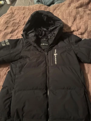 Svart dunjacka Sail Racing med huva - Svart dunjacka från Sail Racing med huva och dragkedja framtill. Jackan har logga på bröstet och är fylld med Allied Feather + Down (500 fill power, 70/30 dun/fjäder). Snygg, stilren och perfekt för kalla dagar.