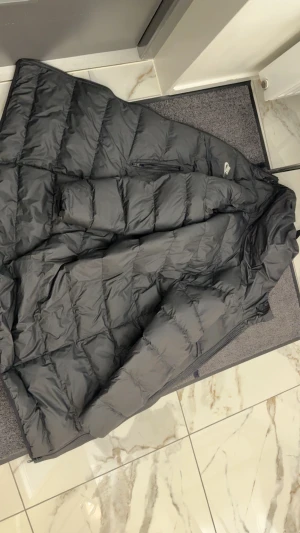 Svart lång dunjacka från Nike - Säljer en svart lång dunjacka från Nike med huva och dragkedja. Jackan har quiltad design och två sidofickor med dragkedja. Perfekt för kalla dagar och har en clean look som funkar till streetwear. Nike-logga på bröstet.